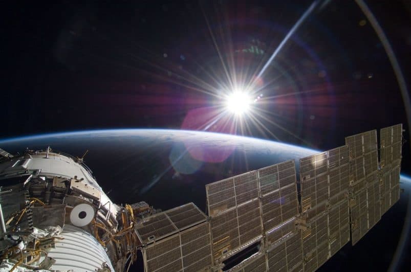 The International Space Station sits in the bottom left corner. In the background, the sun shines over earth.