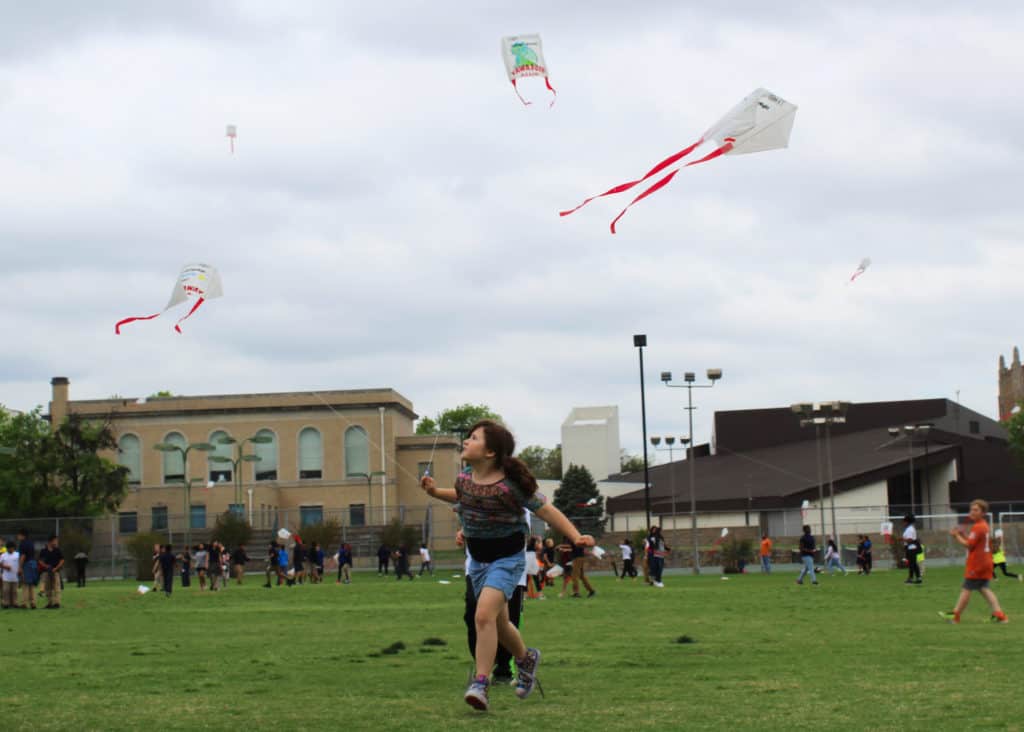 A girl runs with kite in air while students in the background fly kites