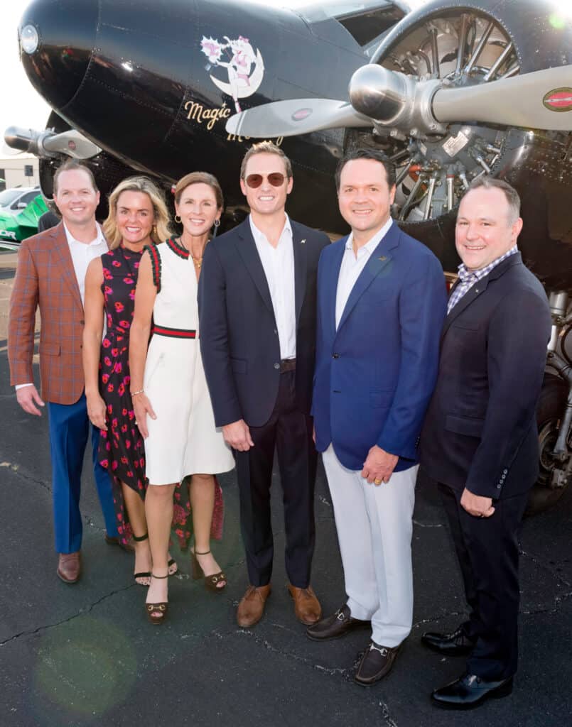 Picture of 5 people standing in front of an old-fashioned medium-sized jet. All are smiling; dressed in business cltohes. 